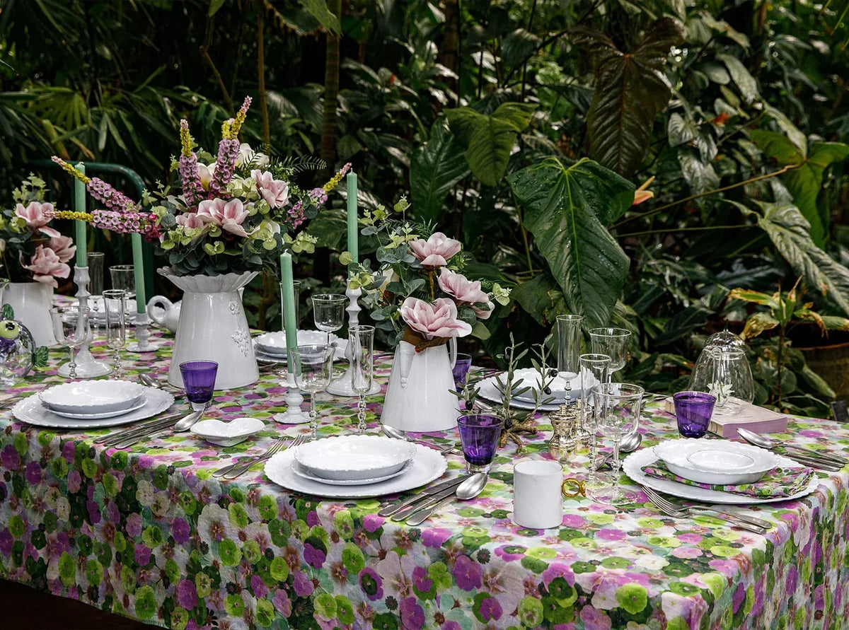 A garden table set with a Summerill & Bishop floral linen tablecloth, white scalloped plates, green taper candles and jugs of pink magnolias