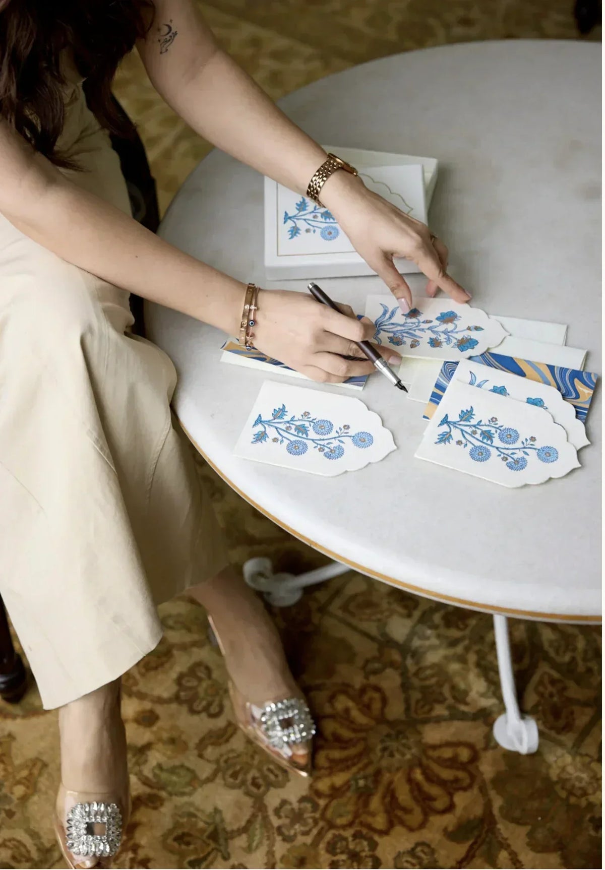 A hand writing on a Peggy & Kate floral note card at a table, pen mid-stroke