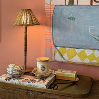 A styled interior scene with a brass table lamp, a candle, a decorative box, and a small book arranged on a tray against a pink wall and abstract artwork.