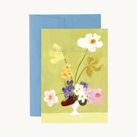 Happy Birthday card of white, pink, purple and yellow flowers in a small white vase on green, with a blue envelope