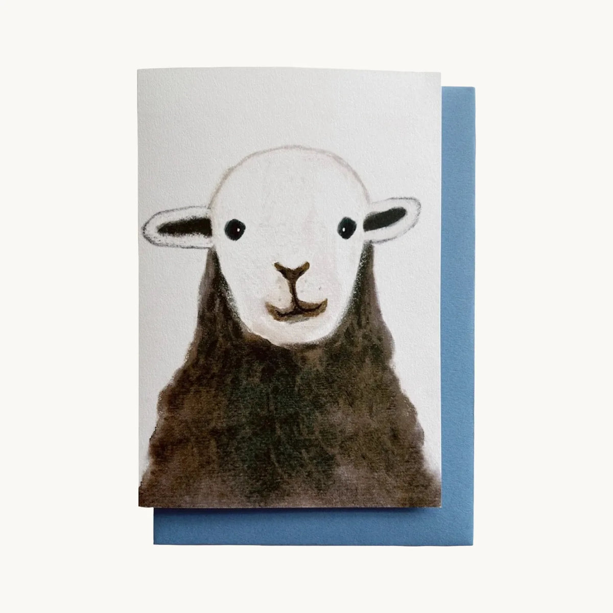 Greeting card of a sheep’s face with a white head and dark brown wool on a white background, with a blue envelope