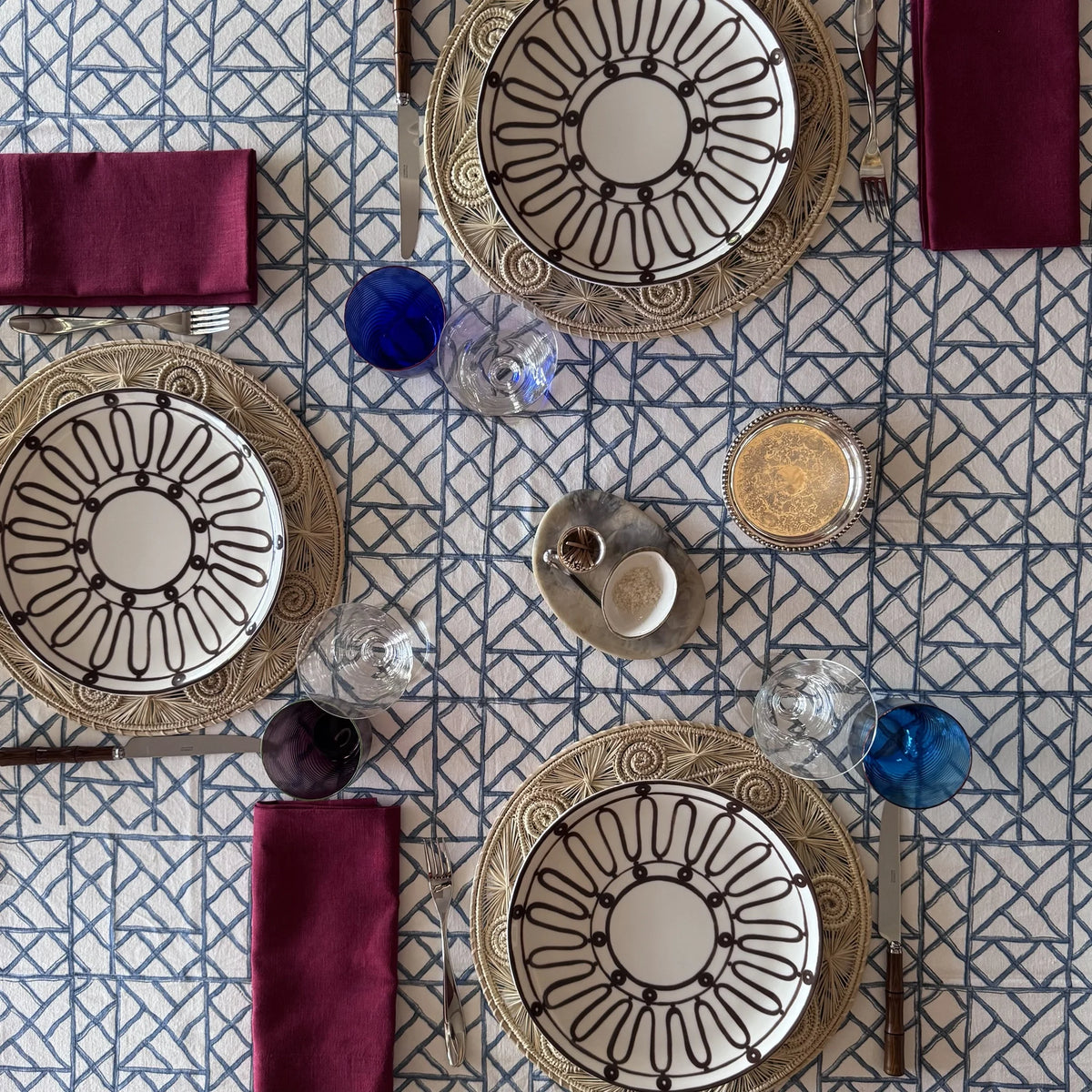 (02) Blue Pagoda block print tablecloth set from above with brown-patterned plates on rattan chargers, burgundy napkins and cobalt glasses