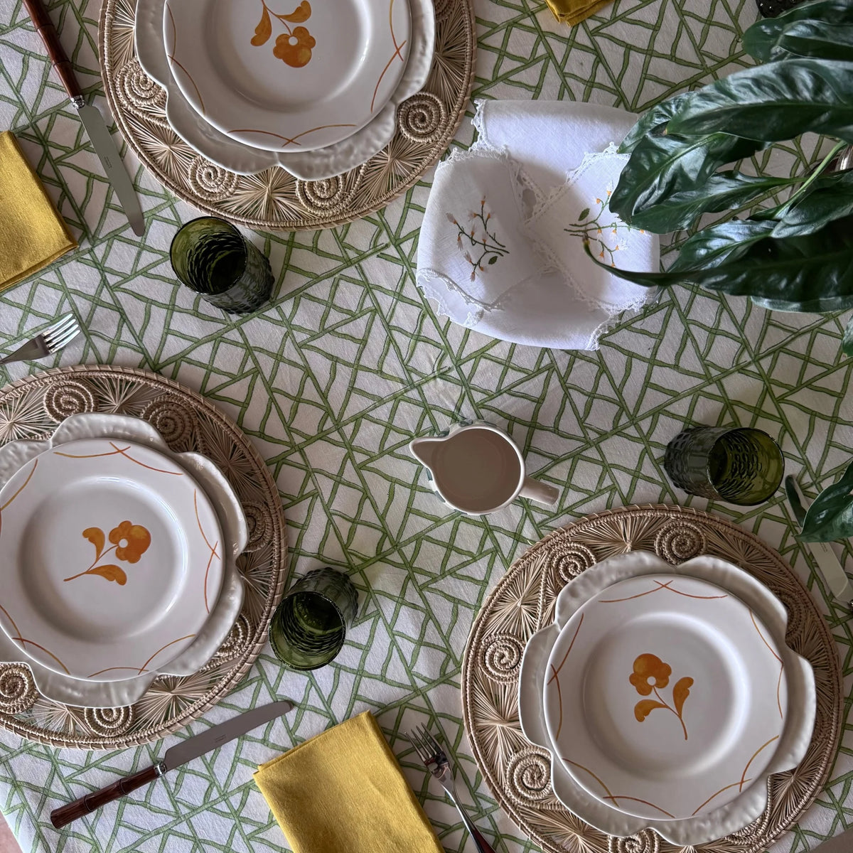 Cane Garden green and white lattice block print tablecloth set from above with rattan chargers, floral plates and mustard napkins