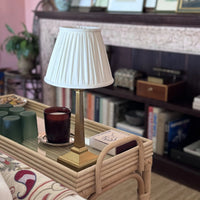 The Voyage Dubai - Cordless Antique Gold Table Lamp with White Pleated Shade - Rechargeable iron table lamp. Styled on a rattan side table in front of a bookshelf.