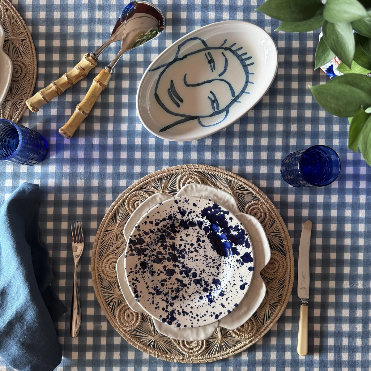 Ischia blue check tablecloth with a line-drawn face oval dish, bamboo-handled servers, navy napkin and blue glass