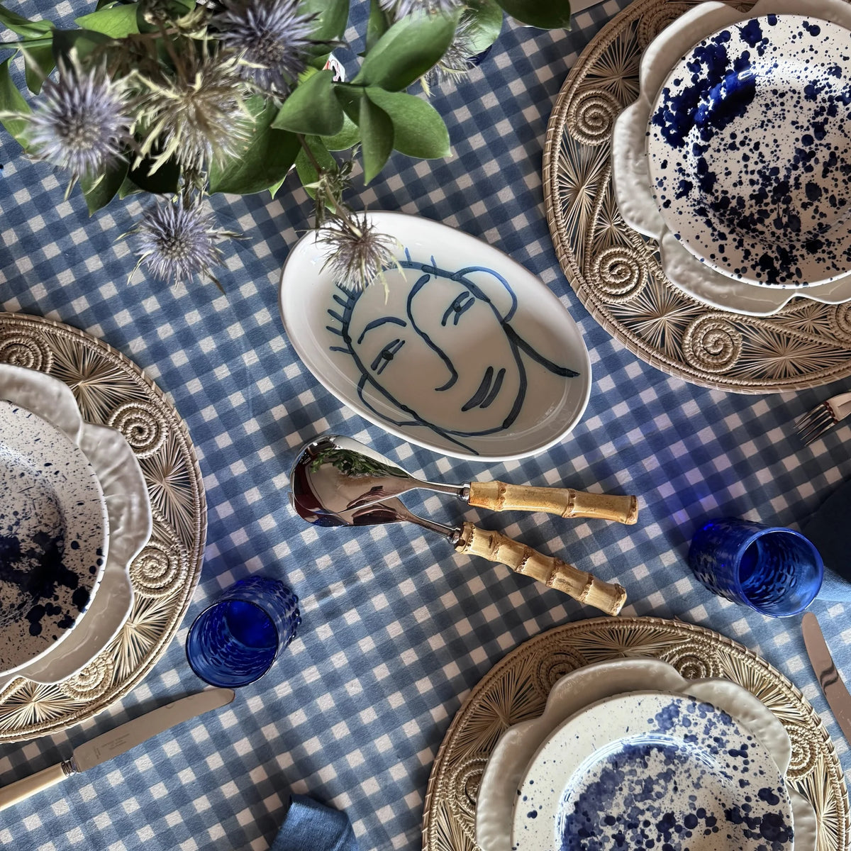 Ischia blue and white check block print tablecloth set from above with rattan chargers, blue splatter plates and thistles
