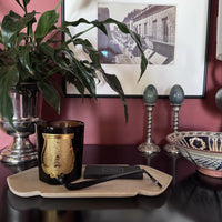 Marble tray on a dark console holding a black glass candle with a gold label and a wick trimmer, with a plant and framed print behind