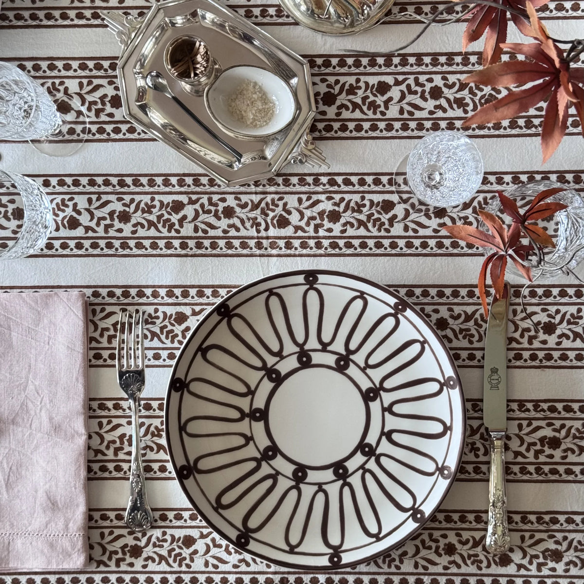 Sienna block print tablecloth in rust-brown floral stripes on cream, set with a patterned plate, silver cutlery and a pink napkin
