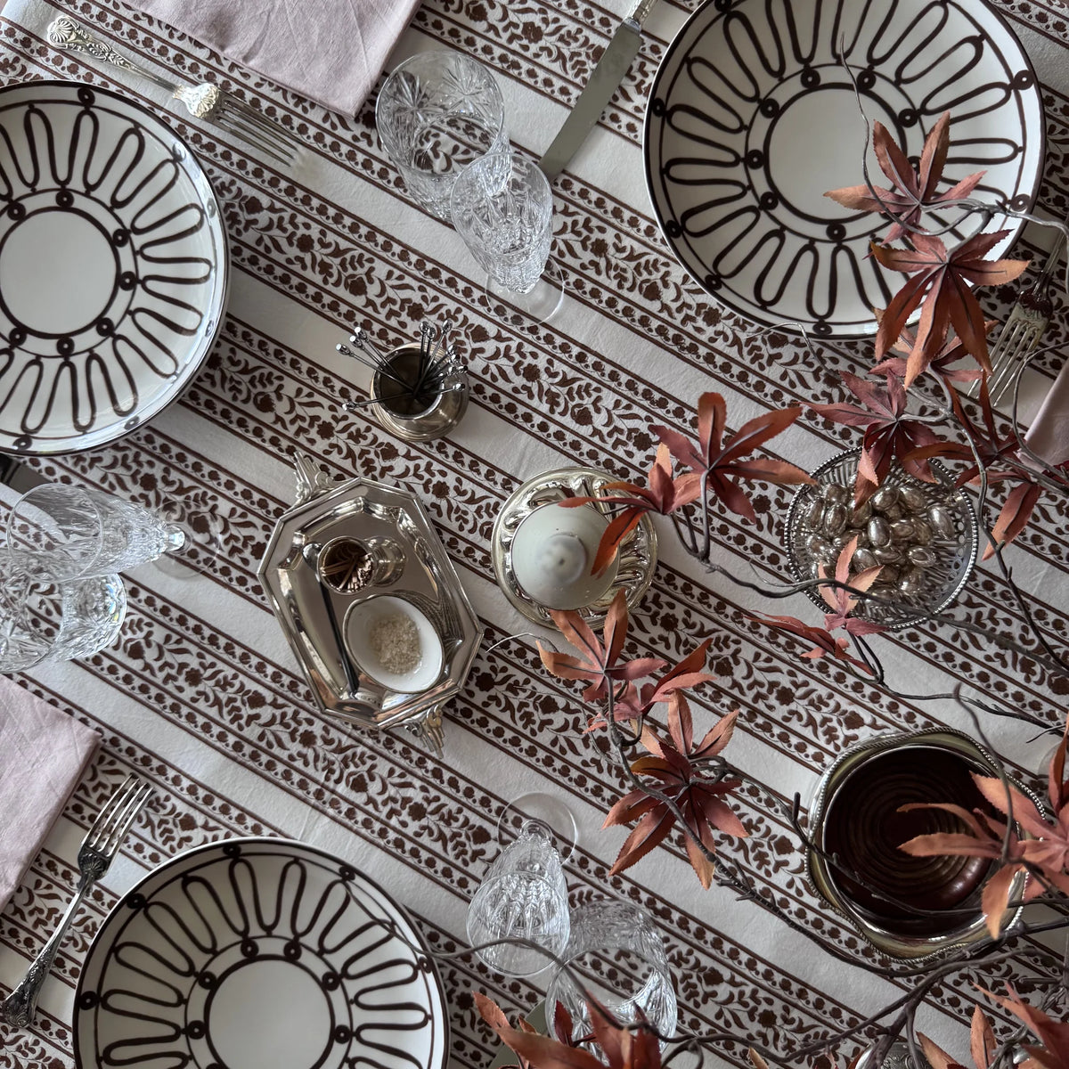 Sienna block print tablecloth in rust-brown floral stripes on cream, set with a patterned plate, silver cutlery and a pink napkin