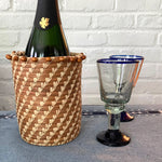 The Voyage Dubai. The perfect accessory for your table!  Handmade and designed by Mayan Hands artisan partners in Sololá, Guatemala this versatile pine needle and raffia basket will accomodate most water and wine bottles. Alternatively, place a glass jar inside to create a unique vase.  Handmade and fair trade.  Origin – Guatemala  Material - pine needles, raffia, wooden beads  Approximate Dimensions: 6" high x 5" diameter